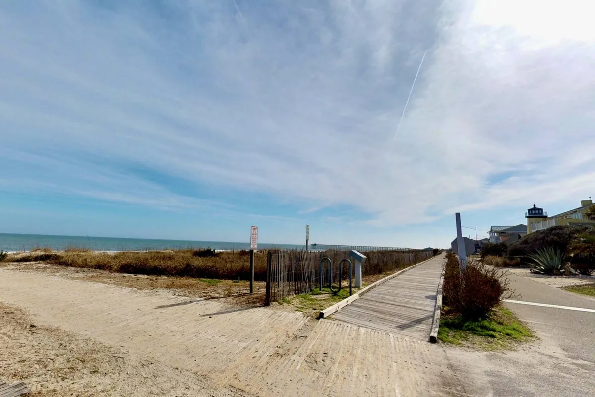 A scenic view of a wooden boardwalk leading over dunes toward the expansive ocean. The foreground features soft sand and native grasses, while a distant lighthouse adds coastal charm. The space is open and inviting, perfect for families heading to the beach for a day of play.