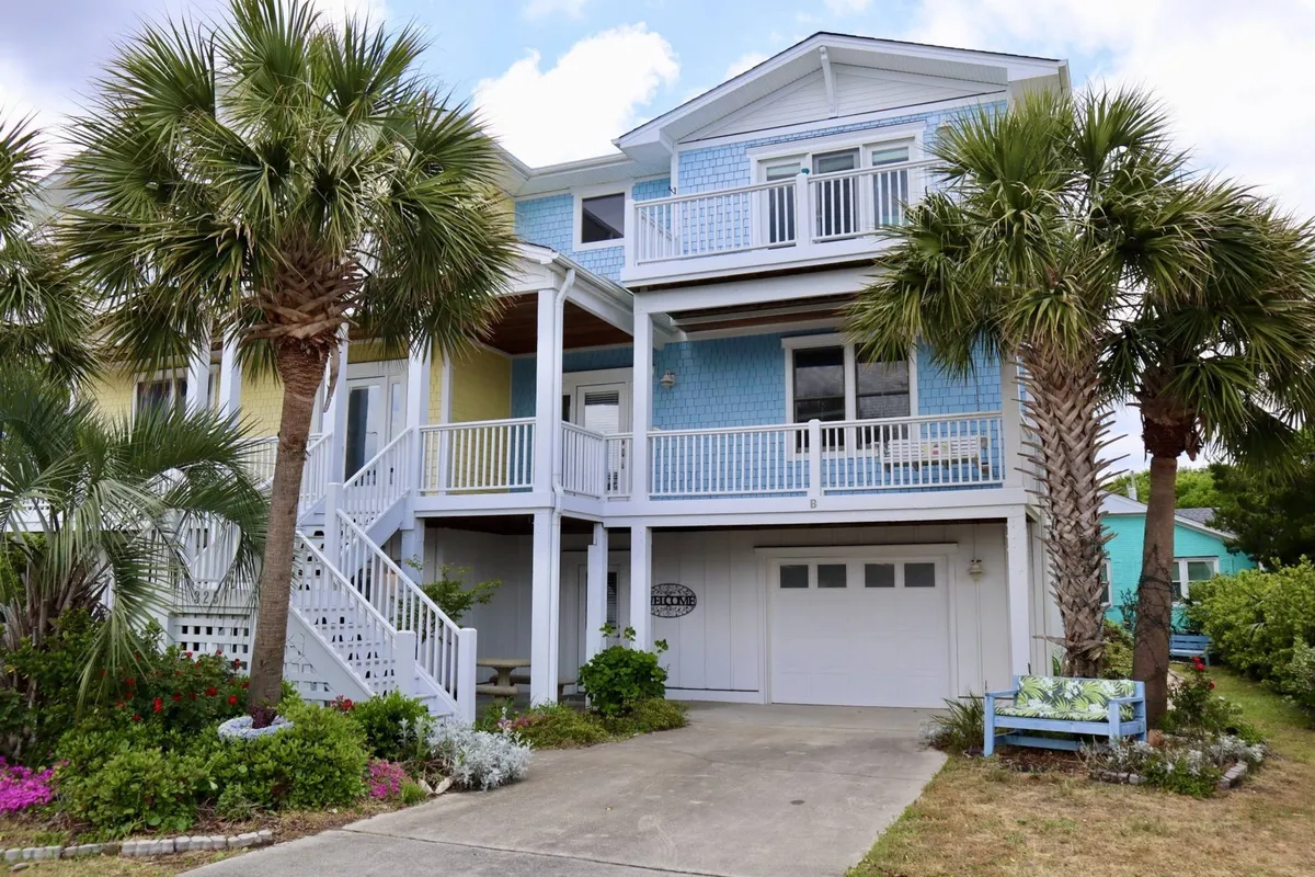 A vibrant, multi-story townhome with a colorful facade, swaying palm trees, inviting front steps, and spacious balconies—set just a few minutes’ stroll from the sandy shores of Kure Beach. The scene radiates coastal charm and hints at leisurely mornings on the porch or sunset gatherings with loved ones.