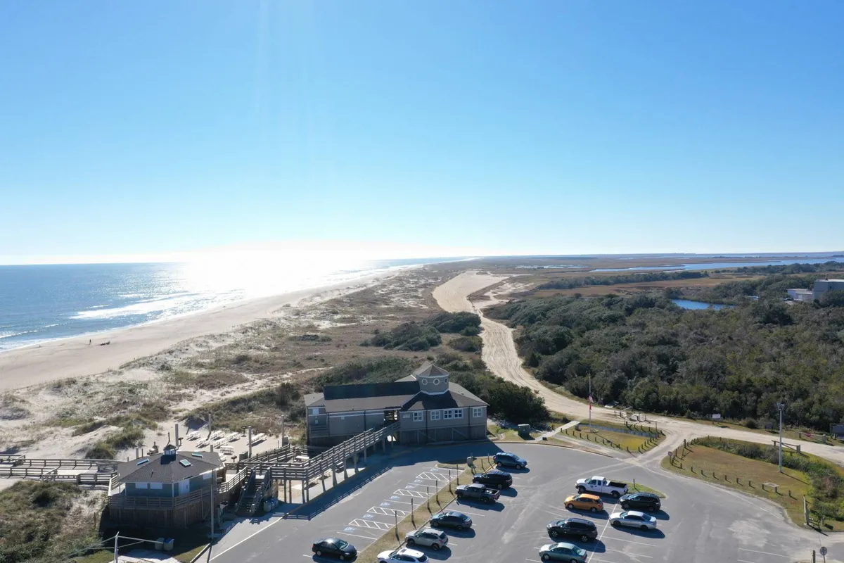 A panoramic view encompassing the ocean, sandy beach, parking area, and the nearby building, all under a wide-open sky. This vantage point radiates a sense of relaxation and possibility for beach gatherings, sunset rides, and nighttime fun.