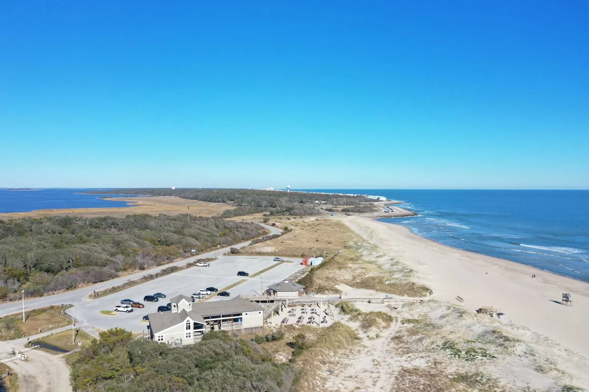 A panoramic exterior view of the beachfront with a coastal path, beachside parking, and the ocean stretching into the distance. The scene is lively yet relaxed, inviting families to swim, beachcomb, picnic, and explore nearby attractions.