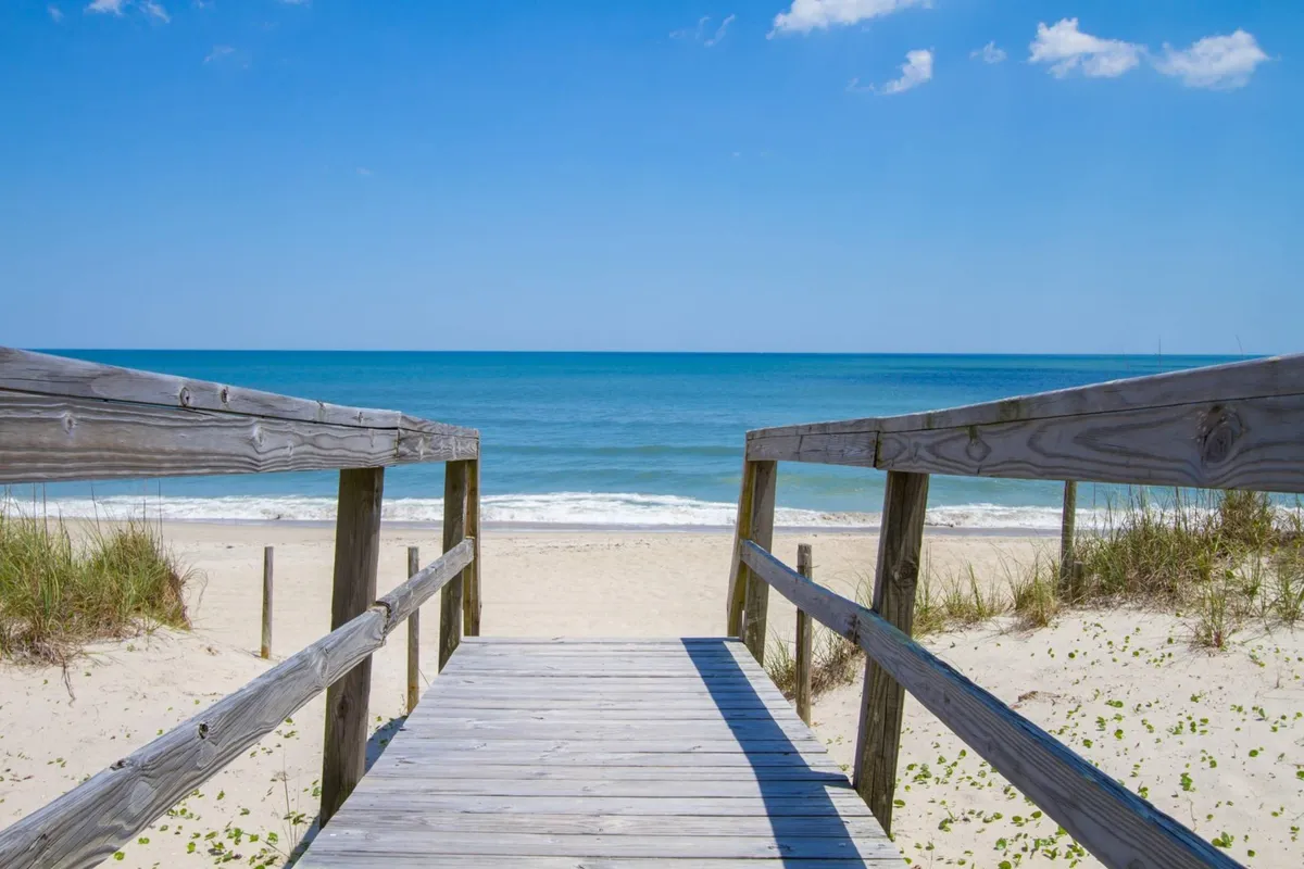 A wide wooden boardwalk leads directly to the sandy beach with the ocean stretching out to the horizon. The scene is serene, with gentle waves, golden sand, and a peaceful sky, evoking a sense of open access and easy movement.