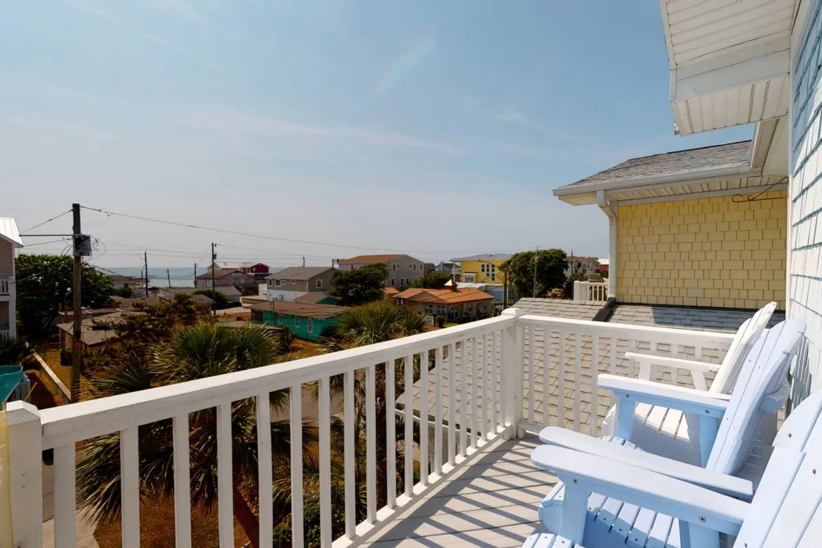 Blue chairs arranged on a balcony face out toward a sweeping ocean view, with colorful coastal homes dotting the background. The scene evokes peaceful mornings and golden-hour relaxation.