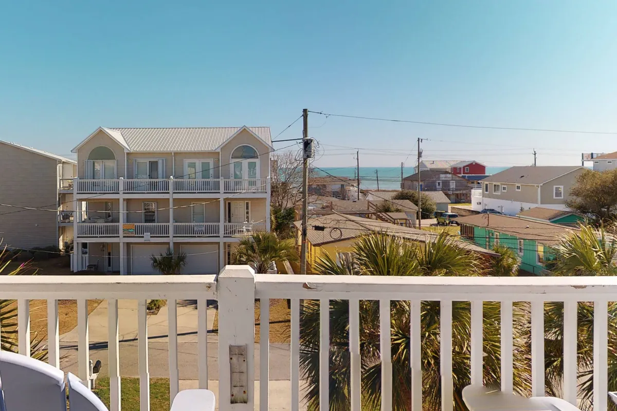 A scenic view from a deck or balcony overlooking the ocean, with a balcony railing in the foreground, palm trees and neighboring houses visible. The setting suggests a tranquil, dog-friendly outdoor space ideal for morning coffee or unwinding with your pup after a day at the beach.