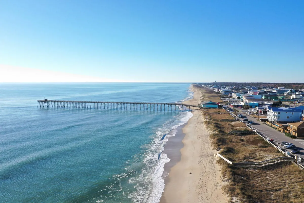 A sweeping view of the Kure Beach Pier stretching out over the ocean, with sandy beach, gentle waves, and nearby beach houses visible. The image captures the energy and allure of the iconic pier—central to both daytime fishing and evening sunset strolls.