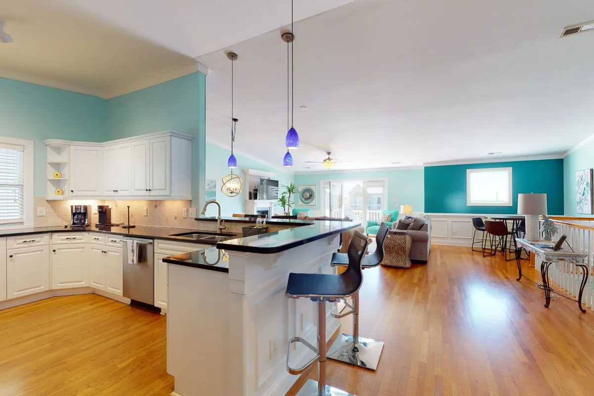 A modern kitchen with a large island and bar stools, seamlessly connected to a living area with expansive windows. The setting invites both cooking and socializing, making it ideal for shared meals and casual gatherings.