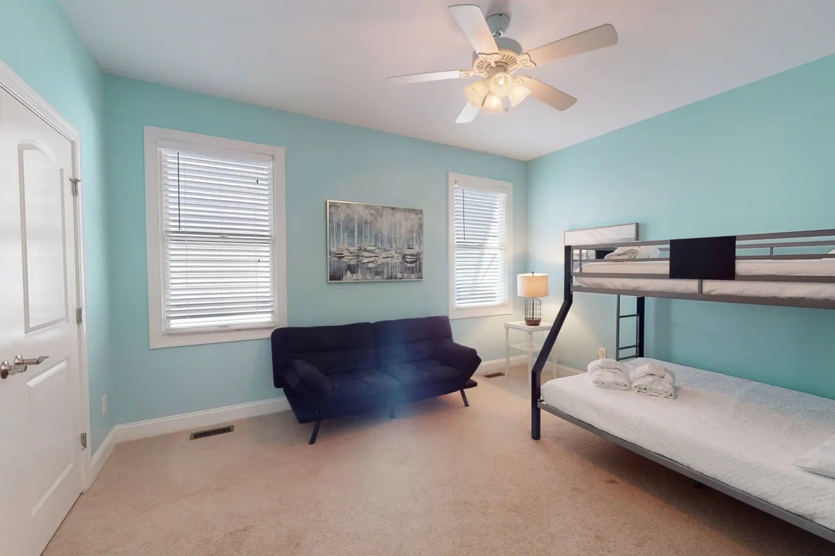 A friendly and versatile bunk/futon room with a bunk bed, a sofa for lounging or reading, playful artwork, and a cozy lamp. The space feels welcoming for children or groups, inviting both sleep and relaxation.