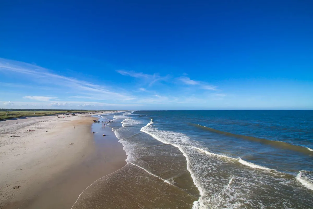A serene shoreline scene with soft ocean waves, a sandy beach stretching into the distance, a blue sky, and people walking along the beach. The landscape is peaceful and spacious, offering a sense of freedom and tranquility.