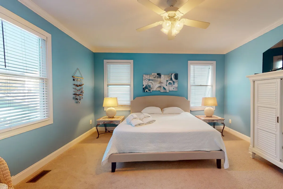 A serene bedroom with a bed featuring white linens and a calming blue accent wall, flanked by bedside tables with lamps and decorative artwork, creating a peaceful environment ideal for rest and recharging.