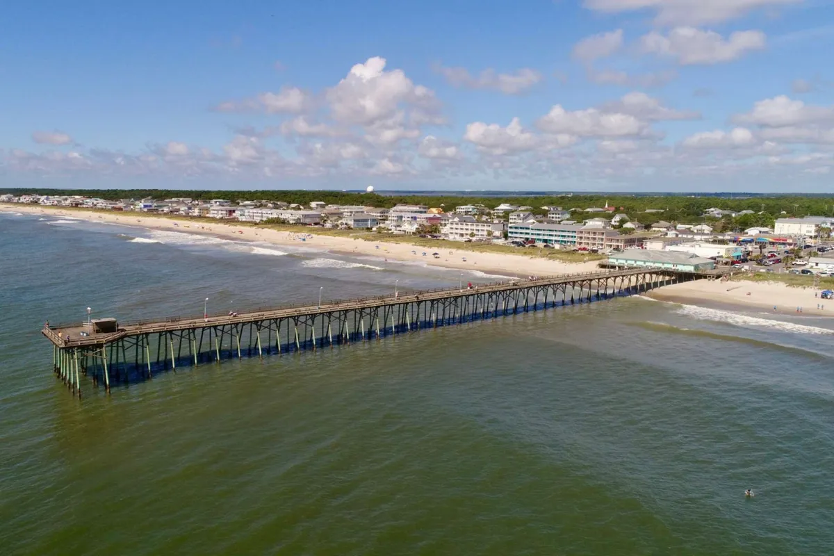 A lively exterior scene of a pier extending over the ocean, beachgoers enjoying the sand, and beachfront houses in the distance. People are visible walking and relaxing on the beach, capturing the energy and easygoing lifestyle of Kure Beach.