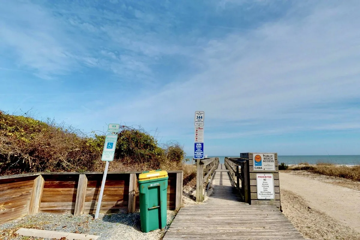 An exterior scene showing a wooden boardwalk leading directly to the ocean, with public access signs and a relaxed beachside atmosphere. The sky is bright and the ocean shimmers in the distance, promising easy beach adventures.