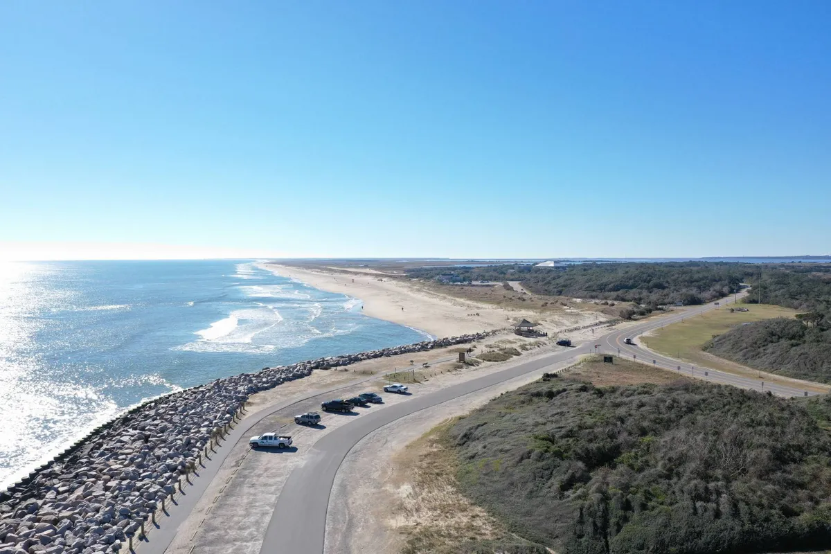 A panoramic view of the shoreline with gentle ocean waves rolling onto the sandy beach, vehicles parked along the roadside, breakwater rocks, and open space for people to walk or explore. There are hints of human activity, such as people walking along the beach and the inviting natural landscape.