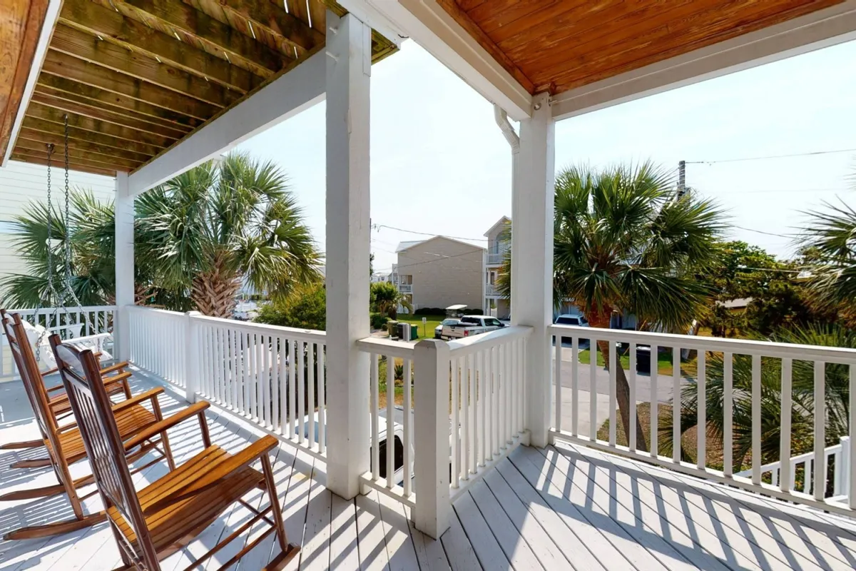 A cozy porch features classic rocking chairs facing out toward palm trees and a deck railing, inviting guests to relax and soak in the ocean breeze.