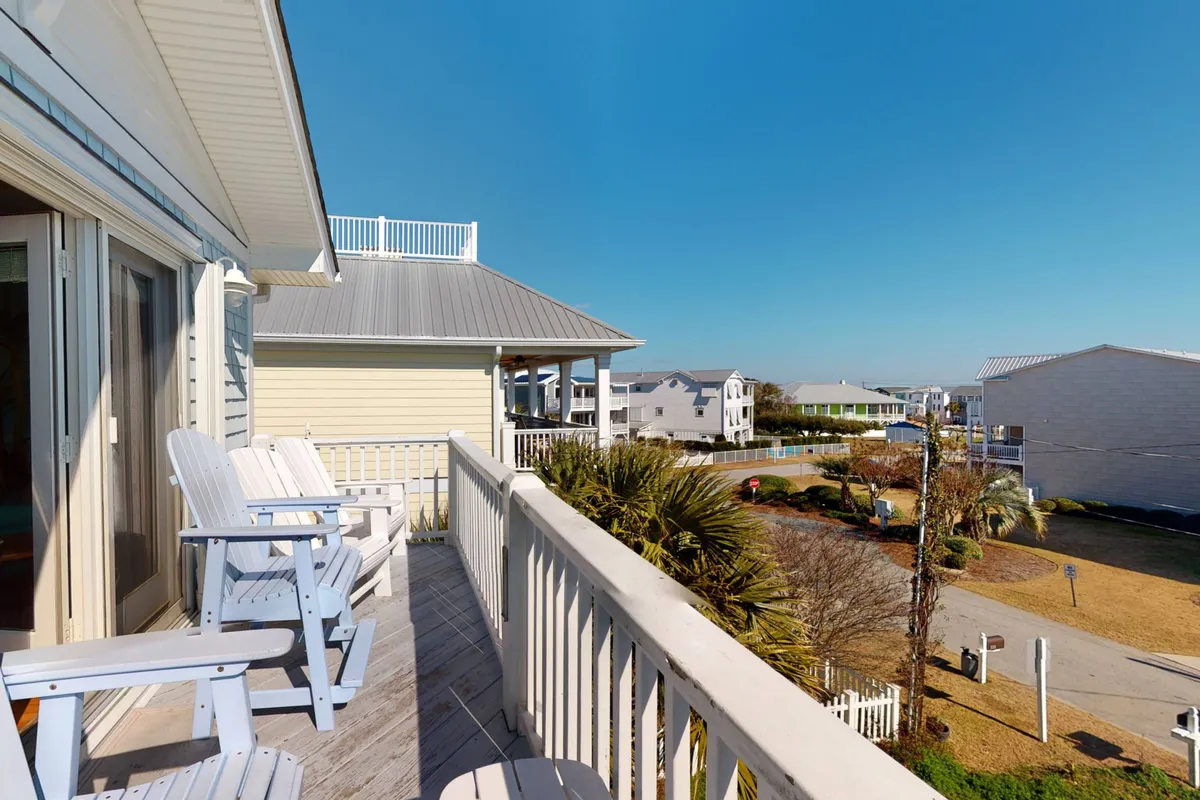 A spacious balcony with comfortable seating overlooks swaying palm trees and neighboring beach houses. The view extends toward the distant ocean, and the setting is clearly designed for relaxation, socializing, and enjoying the coastal breeze.