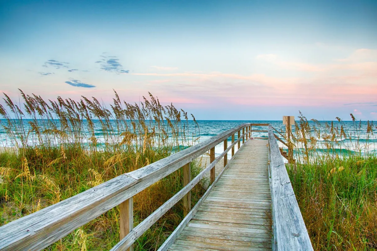 A tranquil wooden boardwalk extends over dune grass toward the rolling ocean, with a glowing sunset sky above. The setting is peaceful and welcoming, highlighting the natural beauty of the coastline and the inviting atmosphere for leisurely strolls.