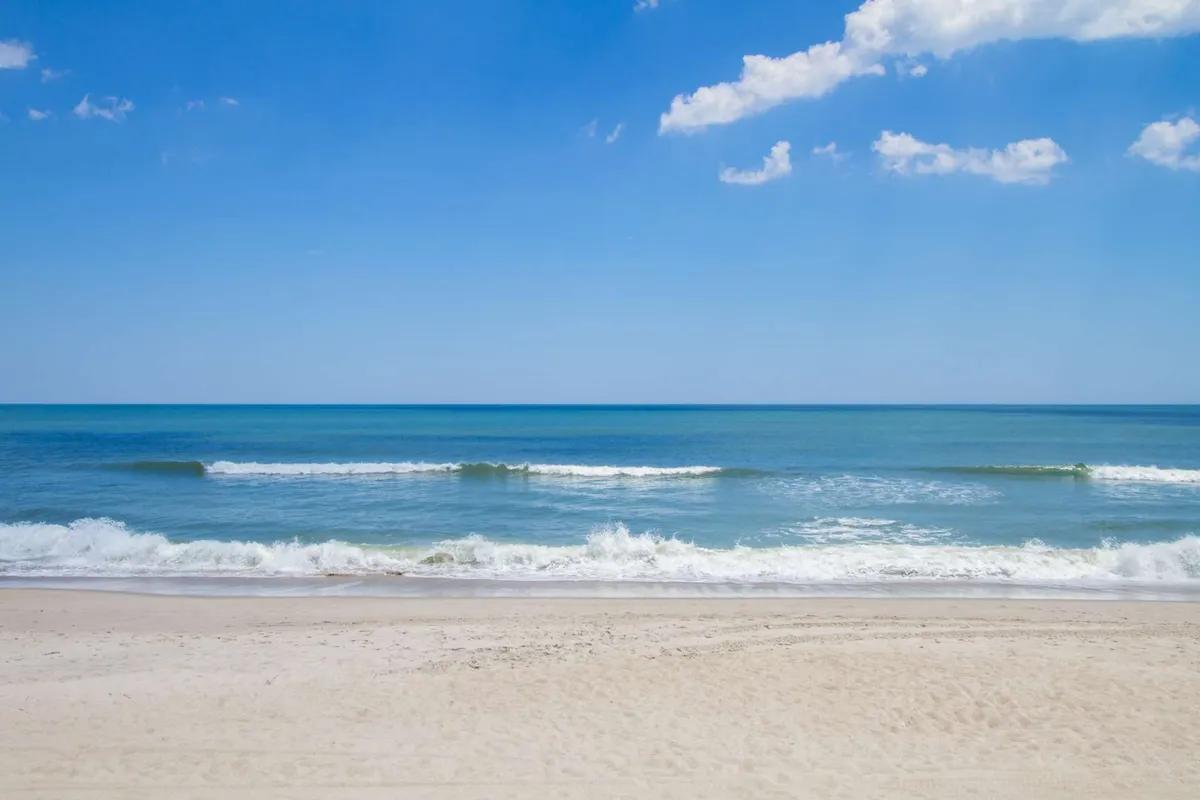 A broad stretch of sandy beach meets gently rolling ocean waves under a clear blue sky. Children’s sandcastles, footprints, and playful activity are implied by the inviting, open expanse—an ideal scene for families gathering, playing, and enjoying the water together.