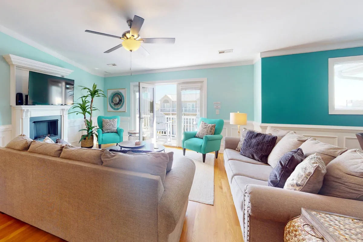 A spacious, light-filled living room featuring a plush sofa set, a cozy fireplace, sliding glass doors opening to a balcony, decorative plants, and accent chairs. The atmosphere is inviting, with ample space for gatherings and relaxation.