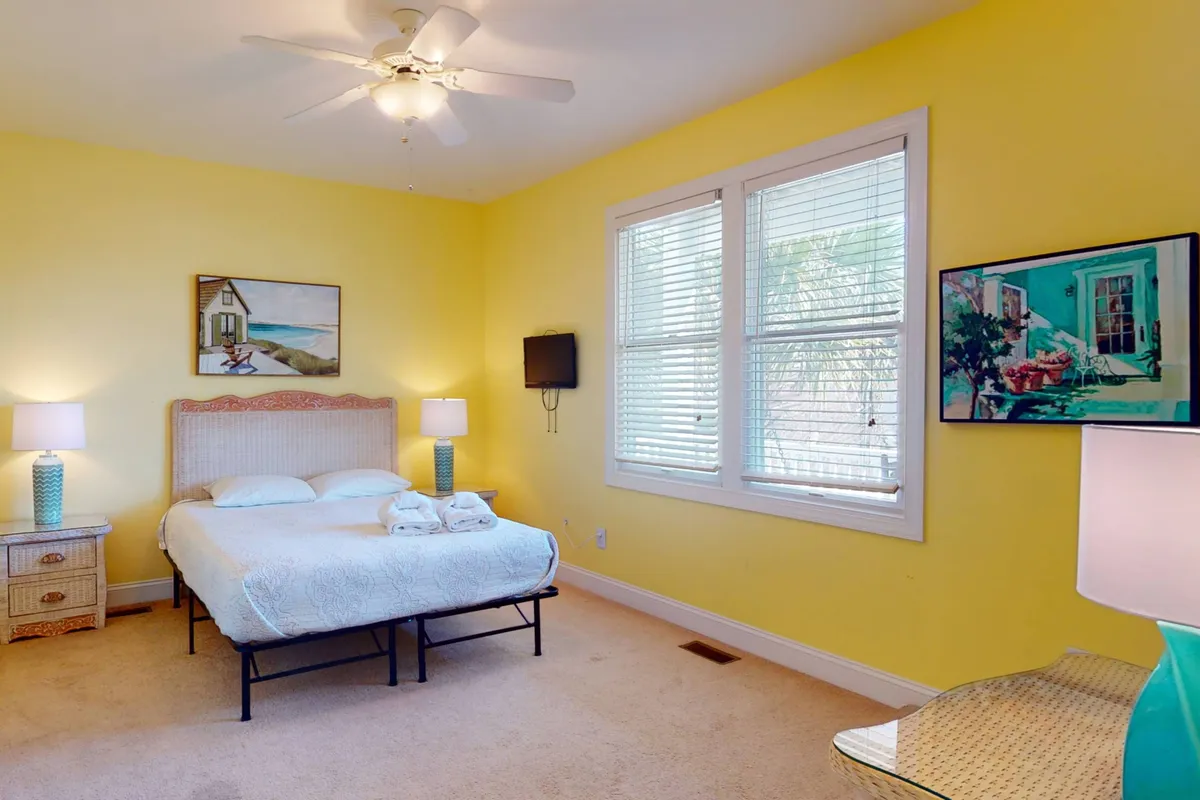 A light and airy bedroom with a bed dressed in crisp white linens, blue lamps on bedside tables, cheerful artwork, and a window letting in natural light. The simplicity and brightness create a peaceful, inviting vibe.